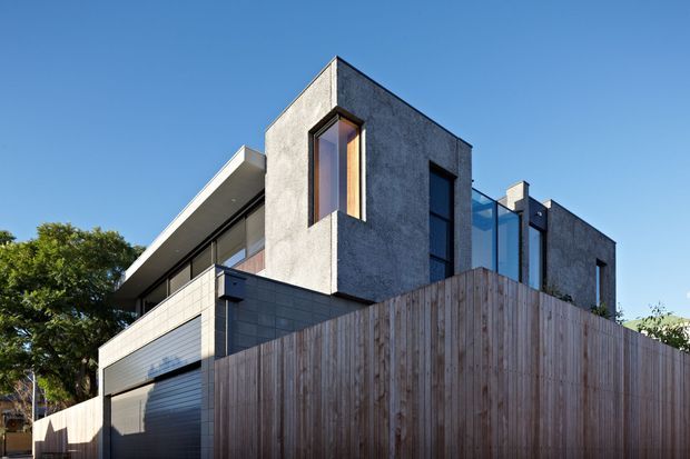 Exterior photograph of a contemporary multi-level residence viewed from the street, featuring a sculptural angular form, dark textured rendered walls, narrow vertical windows, extensive glazing, a projecting flat roof, and a tall vertical timber privacy fence. A garage door and balcony or upper-level terrace are visible, with clear blue sky and surrounding trees in the background.