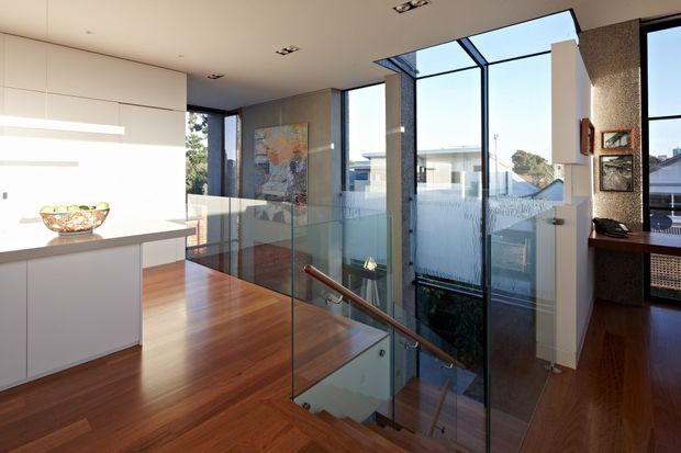 Bright contemporary interior showing a minimalist white kitchen at the edge of a stair landing. Warm timber flooring continues down the staircase, which has a clear glass balustrade and timber handrail. Floor-to-ceiling glazing brings in abundant daylight and overlooks neighbouring buildings. White built-in cabinetry, recessed ceiling lights, and a textured dark feature wall with framed artwork complete the refined space.