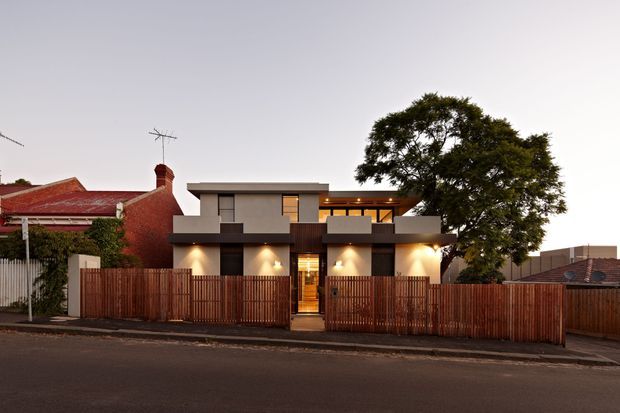 Street-facing photograph of a contemporary two-storey urban residence with a flat-roofed, box-like form, dark and light rendered facade elements, warm exterior lighting, timber privacy fencing, and upper-level glazing. Older brick and tiled-roof homes flank the property, while a mature tree partially frames the right side.