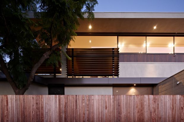 Twilight view of a contemporary multi-level residential facade, featuring a flat roof, broad upper-level glazing, dark horizontal privacy screening, a balcony with metal balustrade, smooth rendered wall surfaces, and a timber privacy fence in the foreground. A mature tree partially screens the building.