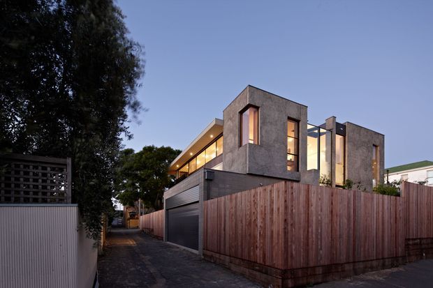 Contemporary multi-storey urban residence viewed from a narrow side lane at dusk. The home features a textured grey rendered or concrete-like facade, strong cubic volumes, tall vertical glazing, warm interior lighting, a projecting upper-level form, and a dark garage door. A tall timber privacy fence encloses the front or side boundary, with mature trees and neighbouring buildings visible around the site.