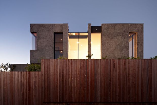 Front exterior view of a contemporary two-storey residence at dusk, largely concealed behind a tall vertical timber boundary fence. The building has strongly geometric concrete-textured wall volumes, narrow vertical windows, and a warm-lit glazed central section creating a recessed entry or courtyard-like void.