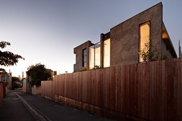 A contemporary multi-level residential building viewed from a narrow urban lane at dusk. The facade combines dark textured masonry or rendered walls with tall warm-lit windows and projecting angular forms. A tall vertical timber privacy fence and masonry base dominate the foreground, with neighbouring urban buildings and vegetation receding along the lane.
