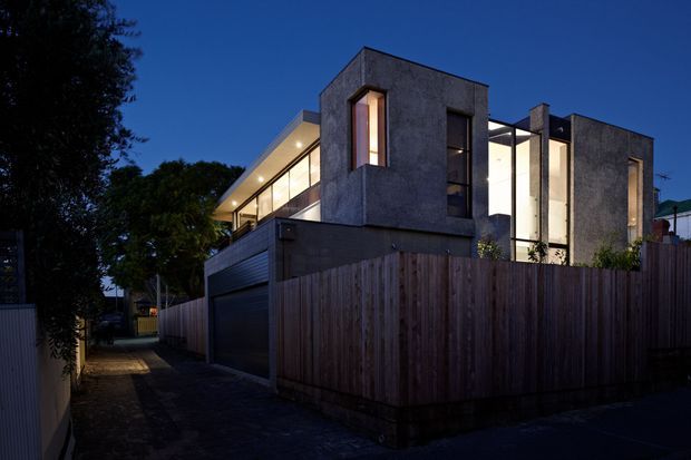 Twilight exterior view of a contemporary multi-level residence, featuring dark textured rendered walls, strong cubic forms, large vertical and horizontal glazed openings, and warm interior lighting. A tall timber privacy fence and narrow paved side access frame the property, with mature trees and neighbouring buildings in the background.