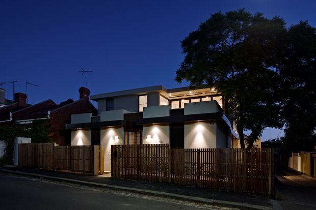 Twilight exterior view of a contemporary multi-level residential building in an urban streetscape. The facade combines dark horizontal cladding and pale rendered surfaces, with recessed illuminated entry or garage bays, upper-level glazing, projecting balconies, and timber slat privacy fencing. Traditional brick homes are visible adjoining the modern residence, while a large mature tree frames the right side.