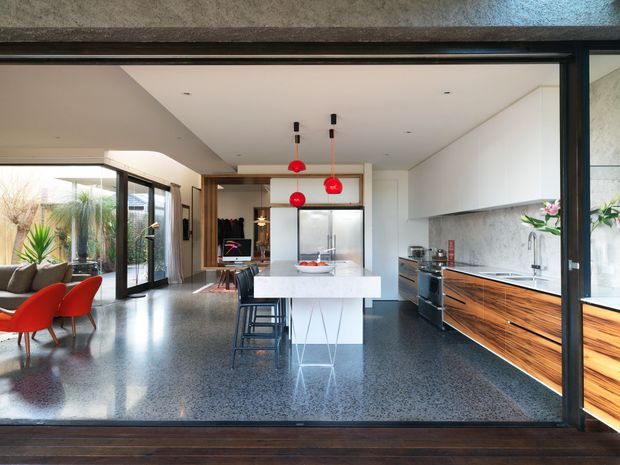 Open-plan contemporary kitchen and dining area viewed through a large black-framed glazed opening. The space features a white island with bar seating, dark polished terrazzo flooring, extensive timber cabinetry, a pale stone or concrete-look splashback, white overhead joinery, red pendant lights, and an adjacent dining and living area with floor-to-ceiling glazing.