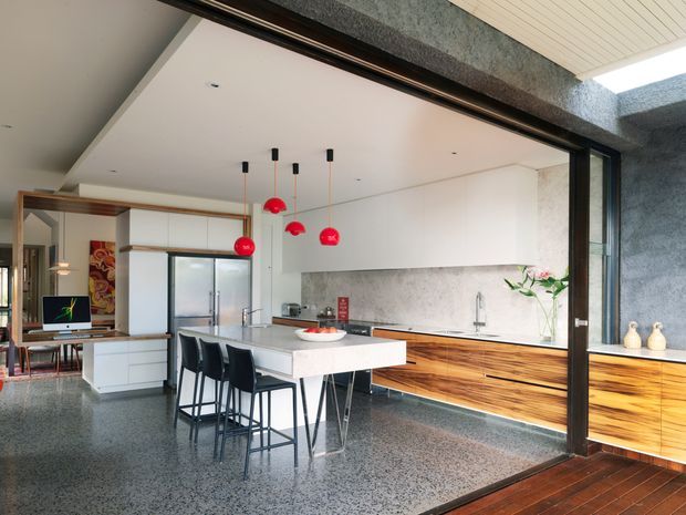 A bright, open-plan contemporary kitchen and dining area viewed through large black-framed glazing. The room features a long timber-fronted kitchen with white benchtops, a central white island with black bar stools, integrated stainless-steel appliances, a textured grey splashback, polished terrazzo flooring, and distinctive red pendant lights. A living area and home office nook are visible beyond the kitchen.