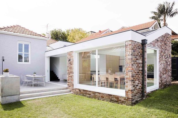 Bright rear exterior view of a renovated residential property, showing a contemporary glazed extension with exposed recycled brick walls and a flat roof. The extension contains a light-filled dining area visible through floor-to-ceiling windows and connects to an older white-rendered building with a tiled roof. A paved courtyard and outdoor dining setting sit beside the extension, with lawn and mature planting around the home.
