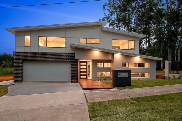 Twilight exterior view of a contemporary two-storey residence with a low-pitched skillion-style roof, light panelled upper facade, dark brick lower walls, integrated garage, illuminated entry, and horizontal windows. The home sits on a suburban landscaped site with a concrete driveway and footpath.