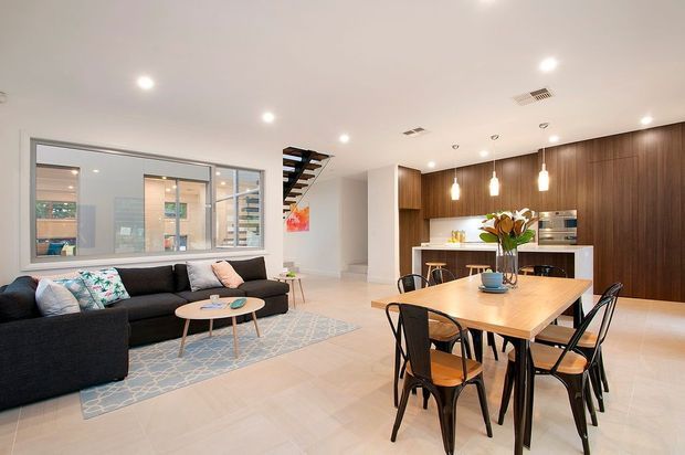 Open-plan contemporary living, dining and kitchen interior with a large dark sectional sofa, timber dining table and black chairs. The kitchen features extensive dark timber cabinetry, a white island, integrated appliances and pendant lighting. A glazed internal partition, staircase and pale tiled flooring contribute to the bright, spacious setting.
