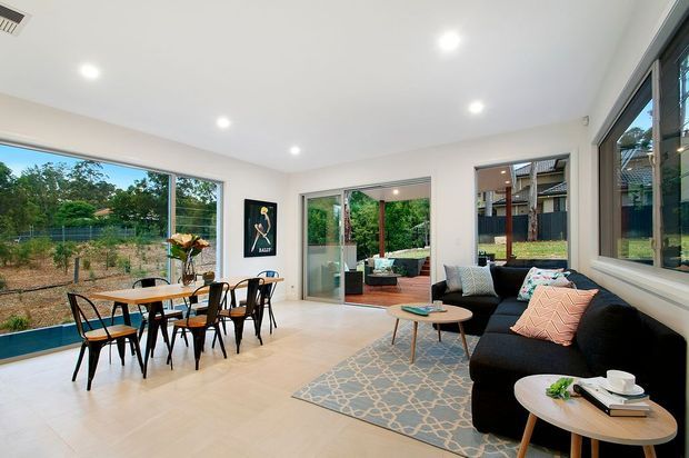 Bright open-plan living and dining room with large corner windows, white walls and ceiling, pale tiled flooring, a timber dining table with black chairs, and a black sectional sofa arranged around nesting coffee tables. Sliding glass doors open to a covered outdoor area, and greenery is visible outside.