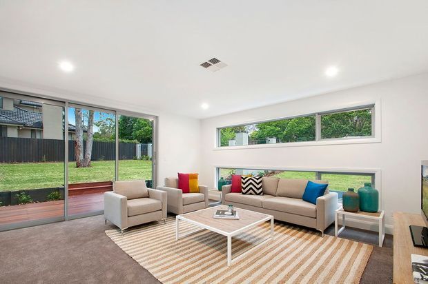 Bright contemporary living room with white walls and ceiling, broad glazed doors and horizontal windows overlooking a landscaped yard, a neutral sofa and armchairs, a striped area rug, and a low white coffee table. Colourful cushions and teal decorative vessels provide small accents.