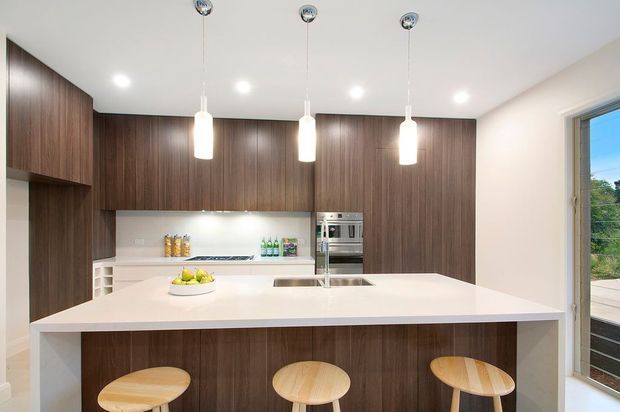 A contemporary kitchen featuring dark timber-grain floor-to-ceiling cabinetry, a large white island with integrated seating, white benchtops, stainless steel appliances, and three pendant lights. A glazed door or window provides views to an outdoor deck.