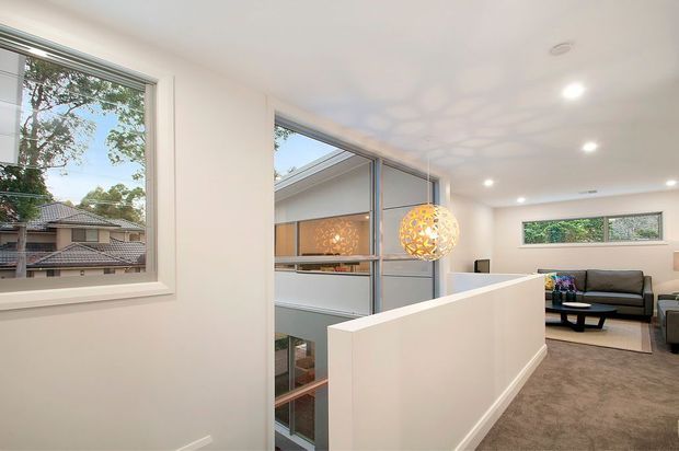Bright contemporary upper-level living area overlooking a lower-level void or stairwell through a glazed partition. The space has white walls, recessed ceiling lights, large windows, grey carpet, a dark sectional sofa, and distinctive spherical pendant lights. A neighbouring residential view is visible through the windows.
