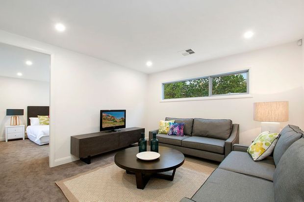 Bright, compact contemporary living room with white walls, recessed ceiling lights, grey sofas, a dark timber media console, round coffee table, and a neutral patterned rug. A large mirror reflects an adjacent bedroom, while a high horizontal window provides greenery views.