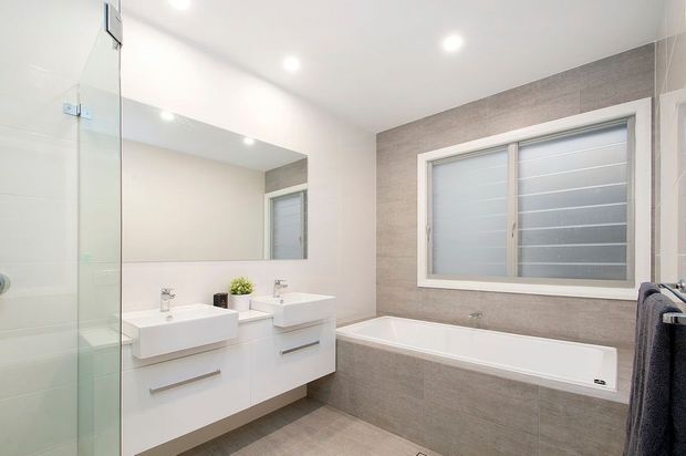 Bright contemporary bathroom featuring a double white vanity with wall-mounted drawers, a large mirror, glass shower enclosure, full-width bathtub, and a horizontal louvre-style window. Neutral grey-beige tiled finishes and recessed ceiling lights create a clean, functional interior.