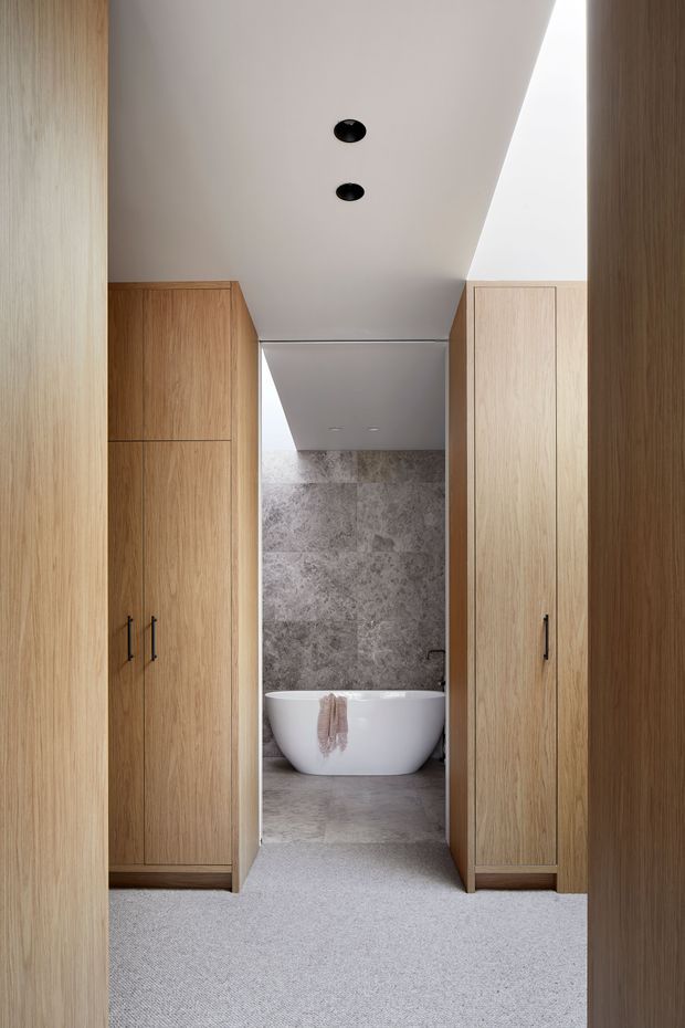 A minimalist interior view through a narrow passage lined with tall light-oak built-in wardrobes, leading to a bathroom with a freestanding white bathtub. The bathroom has large grey stone-look wall and floor tiles, a white sloped ceiling with recessed black downlights, and soft neutral carpeting in the foreground.