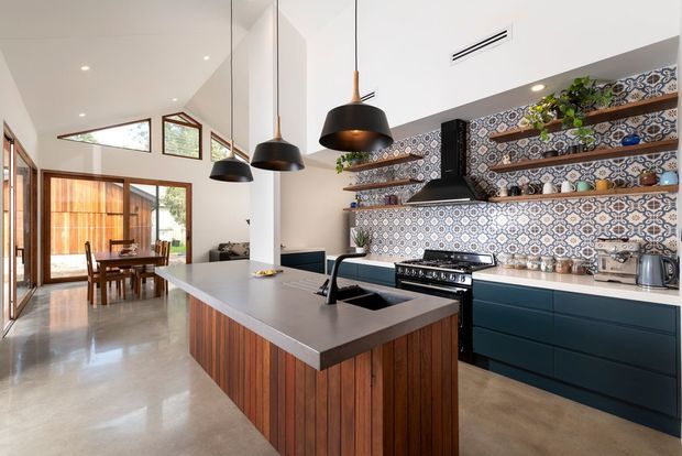 Bright contemporary kitchen with a large island featuring a grey stone benchtop, integrated sink and timber slatted base. Deep blue lower cabinetry, open timber shelves, black pendant lights and a patterned geometric tile splashback define the space. Timber-framed glazing connects the kitchen to a dining area and courtyard beyond.
