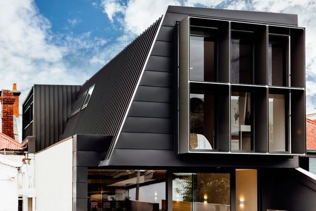 Exterior view of a contemporary multi-level home extension with a dramatic steeply sloping dark metal-clad roof and facade. Large black-framed floor-to-ceiling windows reveal a warmly lit interior, while neighbouring heritage-style brick and corrugated-roof buildings are visible around it.