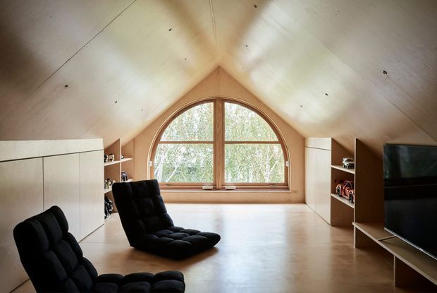 A warm attic-style living space with a dramatic pitched, timber-lined ceiling and a large arched timber-framed window. The room includes black lounge chairs, white built-in cabinetry and shelving, and a television on a low console. Light timber flooring and natural daylight create a calm, minimalist interior.
