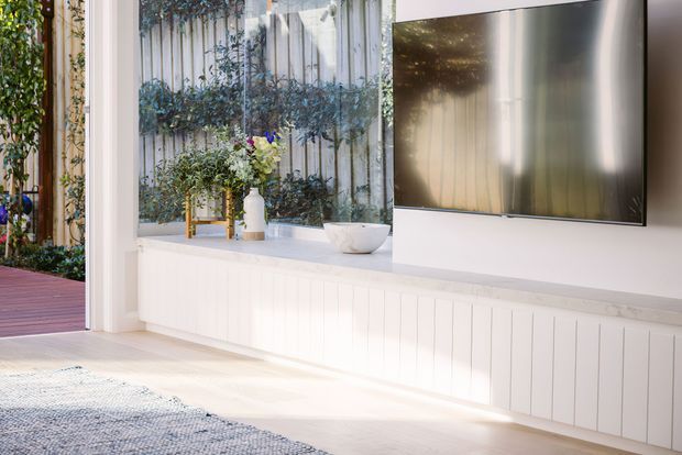 Bright, airy living room with a wall-mounted television above a long white built-in entertainment cabinet featuring vertical panel detailing. Large glazing or an open doorway connects the room to a leafy courtyard or garden, with a timber deck visible outside. The space has pale flooring, white walls and joinery, and understated contemporary furnishings and decor.