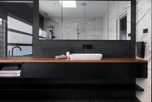 Contemporary bathroom vanity with a white vessel basin set on a warm timber benchtop above matte black floating cabinetry. Large mirrored panels reflect the shower area, while dramatic white marble-look wall surfaces with dark veining and black tiled finishes create a high-contrast, refined interior.