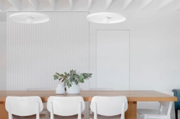 Bright, minimalist dining area with a long natural timber dining table, white molded chairs, and a simple floral arrangement. The room features white vertical wall panelling, exposed white ceiling beams, large circular pendant lights, and a plain white door or wall panel. A partial blue sofa is visible at the right edge.
