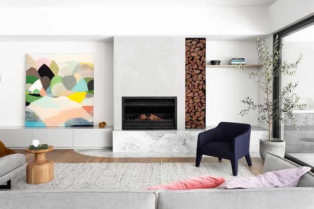 Contemporary living room featuring a white marble fireplace surround, black built-in fireplace, integrated white cabinetry, and a vertical stacked-firewood feature. The room has light timber flooring, a large textured rug, floor-to-ceiling glazing, sculptural furniture, and colourful abstract artwork, creating a refined yet playful interior.