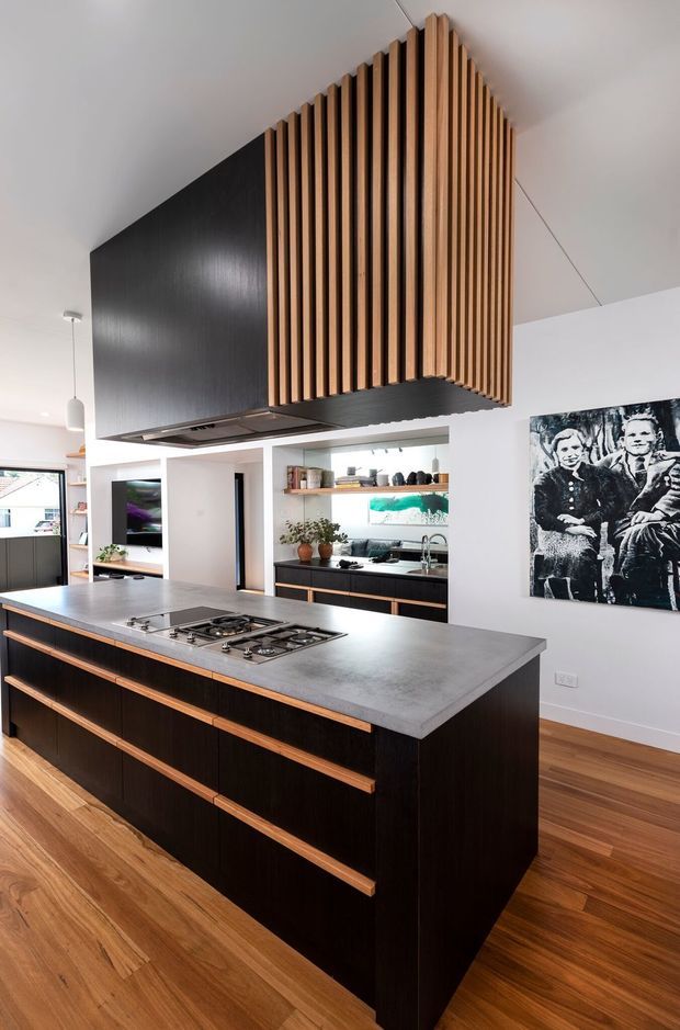 A contemporary kitchen dominated by a large island with a dark timber base, long timber drawer pulls, and a grey stone benchtop containing a gas cooktop. Above the island is a dramatic suspended rangehood enclosure clad in vertical warm timber slats with a dark underside. The kitchen includes dark cabinetry, open shelving, stainless fixtures, white walls, warm timber flooring, and a black-and-white artwork.