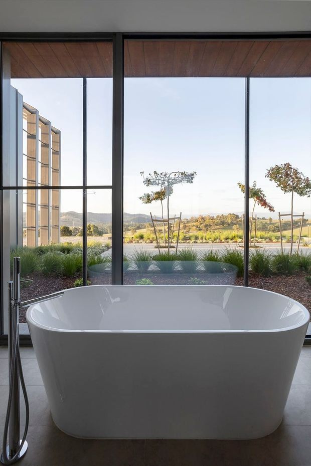 A contemporary bathroom featuring a freestanding white bathtub positioned in front of expansive black-framed floor-to-ceiling glazing. A dark freestanding bath mixer is visible at the left, with large grey floor tiles and a timber-lined ceiling or overhead reveal. Landscaped grounds and rolling hills are visible outside.