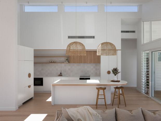 Bright open-plan kitchen and living area with a large white island, integrated white cabinetry, pale tiled splashback, timber flooring, woven pendant lights, and timber bar stools. High-level windows and glazed louvred doors bring in abundant natural light, while a sofa is visible in the foreground.