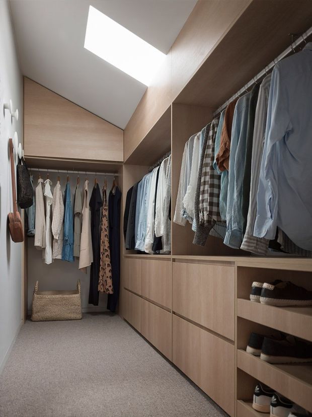 A compact, well-organised walk-in wardrobe with extensive light timber built-in cabinetry, open hanging rails filled with clothing, drawers, and open shoe storage. A large skylight provides natural light, while the carpeted floor and neutral finishes create a calm, practical dressing space.