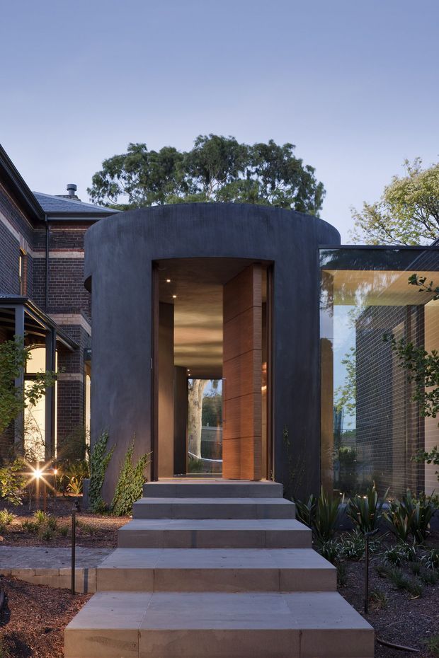 A contemporary residential entrance viewed from the landscaped front approach. Broad concrete steps lead to a tall, dark curved entry pavilion with a recessed timber-clad doorway and warm concealed lighting. Existing dark brick wings and extensive glazing flank the entry, with drought-tolerant planting and mature trees surrounding the path.