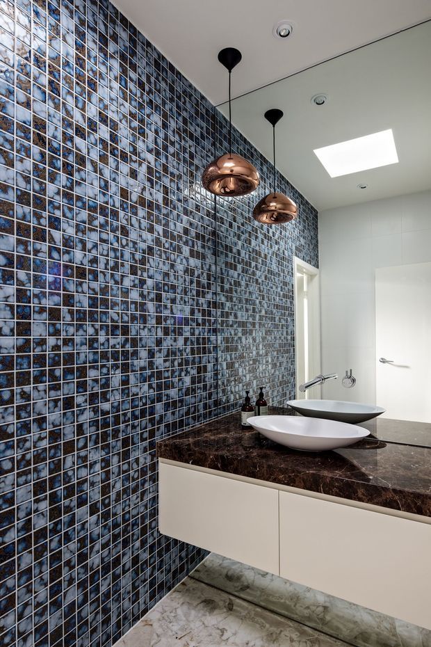 A contemporary bathroom vanity with two white vessel basins set on a dark polished stone benchtop and floating pale cabinetry. One wall is covered in small blue, grey and brown mosaic tiles, contrasted with white tiled walls and a light veined stone floor. Copper pendant lights hang above the vanity, with a large mirror and skylight adding brightness.