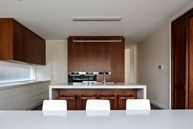 A contemporary kitchen and dining area viewed from the dining table. The room features dark timber cabinetry, integrated appliances, a white island bench with timber bar stools, pale cabinetry along the window wall, and a clean white ceiling and dining surface. A dark timber-framed doorway is visible on the right.