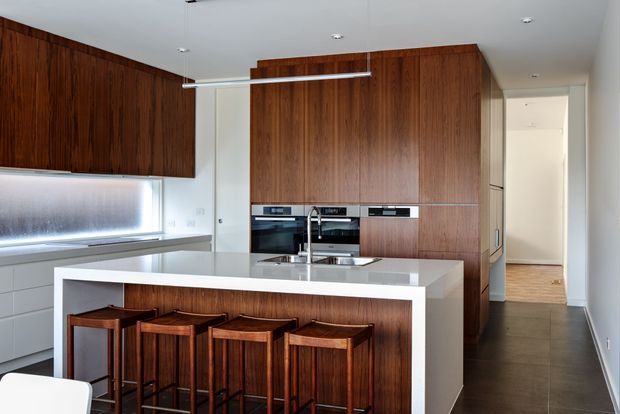 A refined modern kitchen with extensive dark timber veneer cabinetry, integrated stainless-steel appliances, and a large white island with an undermount sink and four timber bar stools. A slim linear pendant hangs above the island, while a hallway extends beyond the kitchen. The palette is warm timber, white, and grey with clean-lined contemporary detailing.