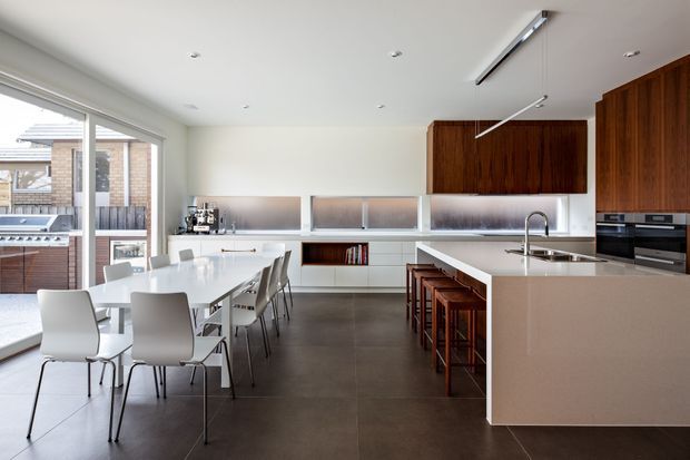 Bright, open-plan contemporary kitchen and dining area with a long white dining table beside full-height glazing. The kitchen features a large pale stone island with integrated sink and seating for several people, dark timber overhead cabinetry, white lower cabinetry, frosted glass-fronted storage, built-in appliances, and dark large-format floor tiles. A barbecue and neighbouring brick building are visible outside.