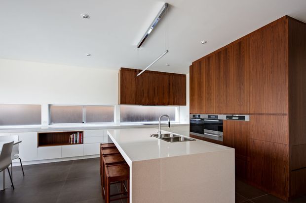 Contemporary kitchen with a large pale stone island, integrated stainless-steel sink and tap, dark timber cabinetry, white lower cabinetry, frosted clerestory-style glazing, and dark tiled flooring. The space includes a compact dining area and streamlined built-in appliances, with a clean, minimalist composition.
