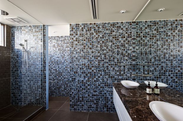Contemporary bathroom featuring a large mosaic-tiled wall and shower enclosure, dark floor tiles, and a double vanity with white vessel basins on a dark stone benchtop. Recessed ceiling lighting and concealed linear air-conditioning vents contribute to the clean, modern fit-out.
