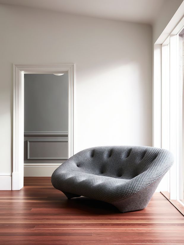 A bright, minimalist interior featuring a sculptural grey tufted lounge chair on rich reddish-brown timber flooring. White walls, detailed traditional door trim, and tall white-framed glazing create a refined contrast between classic architectural elements and contemporary furniture.