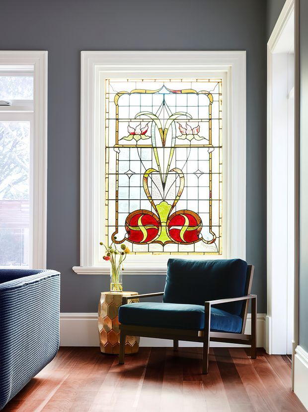 A refined sitting area with a dark teal upholstered armchair positioned in front of a large decorative stained-glass window. The room features charcoal-grey walls, crisp white layered window and door trims, warm timber flooring, and a small metallic gold side table with flowers. A second upholstered piece is partially visible at left, while bright natural light enters through the surrounding windows.