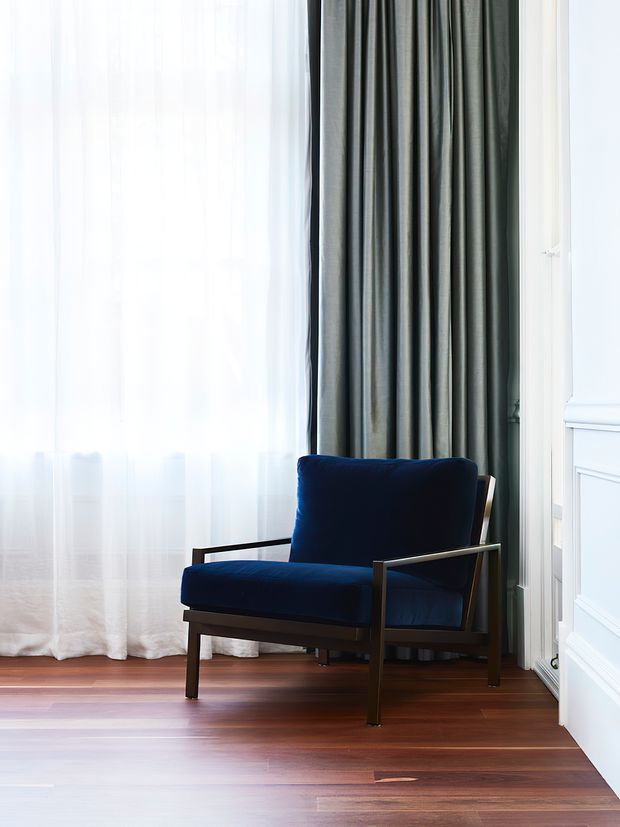 A refined interior corner featuring a navy upholstered lounge chair with slim dark wood or metal arms, positioned beside a tall window. Full-height sheer white curtains are layered with dark grey drapes, while warm timber flooring and white decorative wall panelling create a classic, polished setting.
