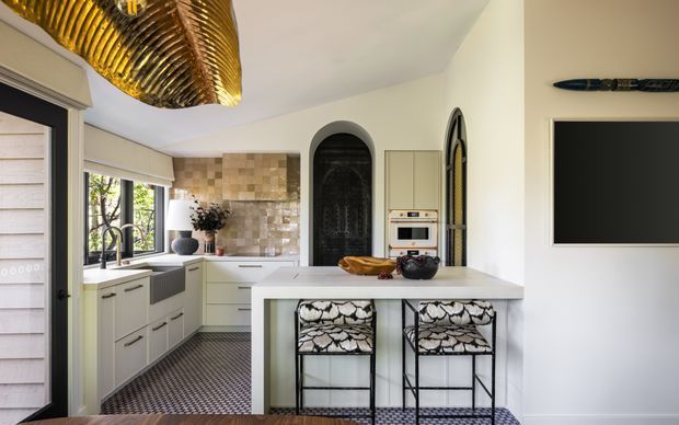 A bright, refined kitchen featuring pale built-in cabinetry, a large central island with two patterned bar stools, a tiled backsplash, arched black-framed openings and doors, integrated appliances, and a dramatic sculptural gold-toned ceiling light. A wall-mounted television is visible at right, with patterned flooring and garden views through the windows.