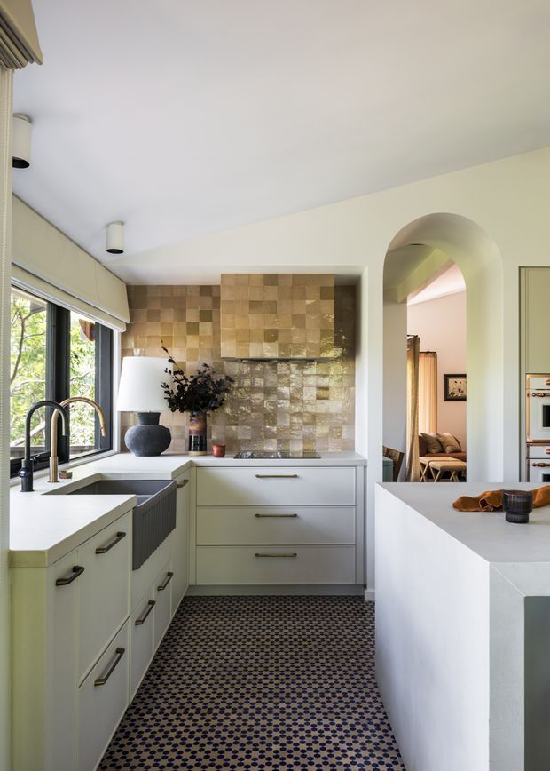 Bright, narrow galley-style kitchen with pale built-in cabinetry, light benchtops, dark hardware, a large window, and a striking metallic textured tile splashback. A patterned dark geometric floor runs through the space, with an adjoining room visible through a rounded archway and a partial second kitchen area at right.