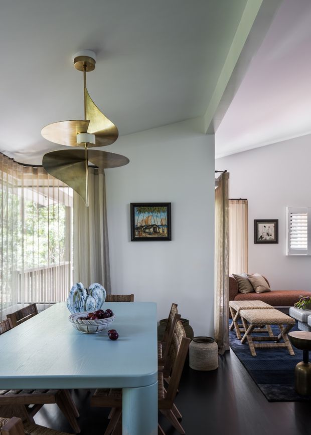 A refined open-plan dining area flowing into a living room. The dining space features a pale blue rectangular table with timber dining chairs, a sculptural brass pendant light, sheer curtains, framed artwork, and dark timber flooring. Through the wide opening, the adjacent living room contains a low sofa, woven ottomans, a dark rug, shutters, and neutral furnishings. The interior has soft daylight, muted pastel walls, and warm metallic and timber accents.