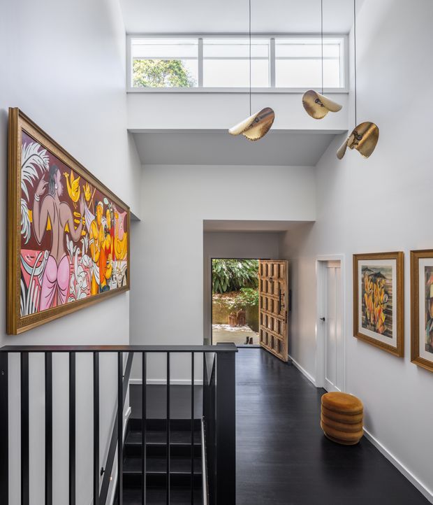 A bright, double-height hallway and stair landing with white walls, dark timber flooring, and a black metal stair balustrade. Framed colourful artwork lines the walls, while sculptural suspended wooden elements hang above the stairwell. A high clerestory window brings in natural light, and an open timber door leads outdoors at the end of the corridor.