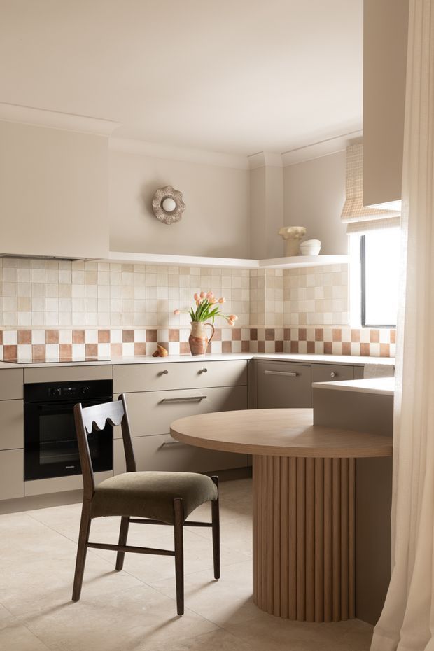 Warm, refined kitchen interior featuring cream cabinetry, a tiled checkerboard backsplash in soft beige and terracotta tones, integrated black oven, pale tiled flooring, and a rounded timber breakfast island with a vertically fluted base. A sculptural upholstered chair, sheer curtains, roller blind, decorative objects, and flowers add a soft, contemporary-classic character. The room is compact to medium-sized with a standard-height ceiling and abundant natural light.