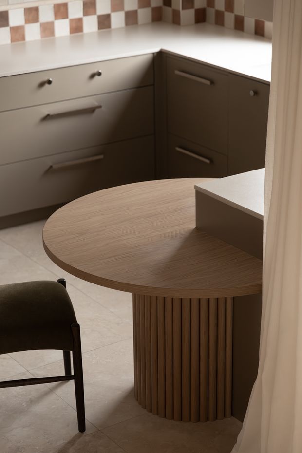 Close-up interior view of a compact, warmly lit kitchen or dining nook. Taupe-grey built-in cabinetry with simple metal handles lines the rear wall beneath a white benchtop and a checkerboard tiled splashback in white and brown tones. In the foreground is a round timber table with a fluted cylindrical pedestal base, beside a dark upholstered chair. Light tiled flooring and a soft curtain complete the scene.