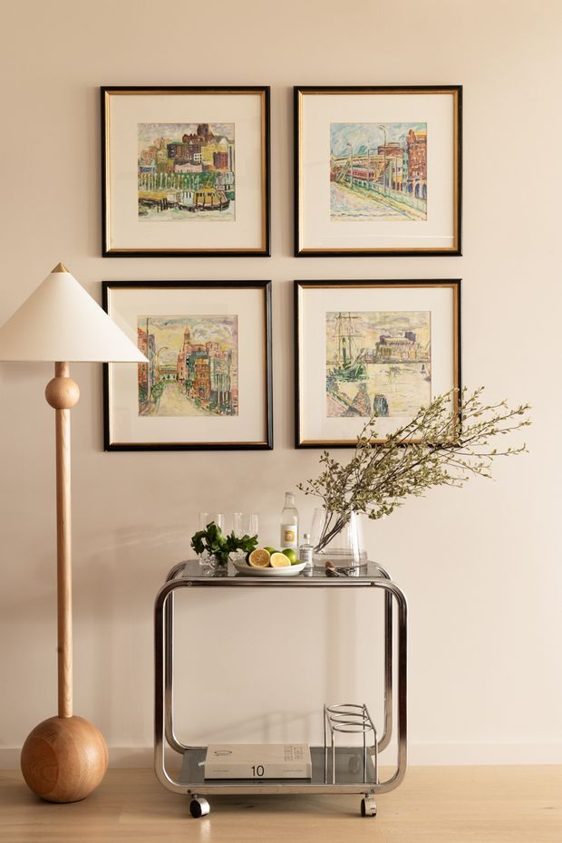 A warmly styled interior vignette featuring a slim metal drinks trolley against a beige wall, topped with glassware, a decanter, citrus and a tall branch arrangement. Four framed urban artworks hang in a neat two-by-two grid above, while a sculptural wooden floor lamp stands to the left. The space has pale timber flooring and a calm, refined mid-century-inspired arrangement.