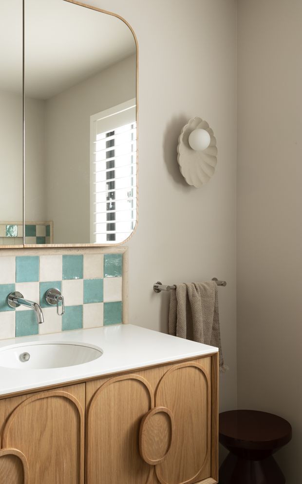 A warmly styled bathroom vanity with a white integrated basin and white benchtop, turquoise and cream checkerboard tile splashback, light timber arched-panel cabinetry, and brushed metal tapware. A large rounded-corner mirror, white plantation shutters, sculptural wall light, towel rail, and dark timber stool complete the compact space.